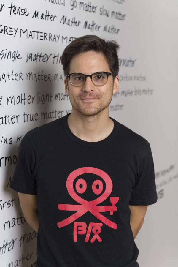 A man wearing glasses and a black t-shirt stands in front of a wall with text written in various scripts repeatedly saying 