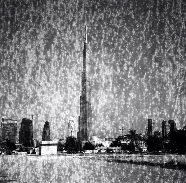 Grainy black and white photograph of a tall skyscraper among much smaller buildings