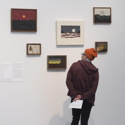 A person wearing a hoodie and beanie faces six small and medium sized paintings on a gallery wall.