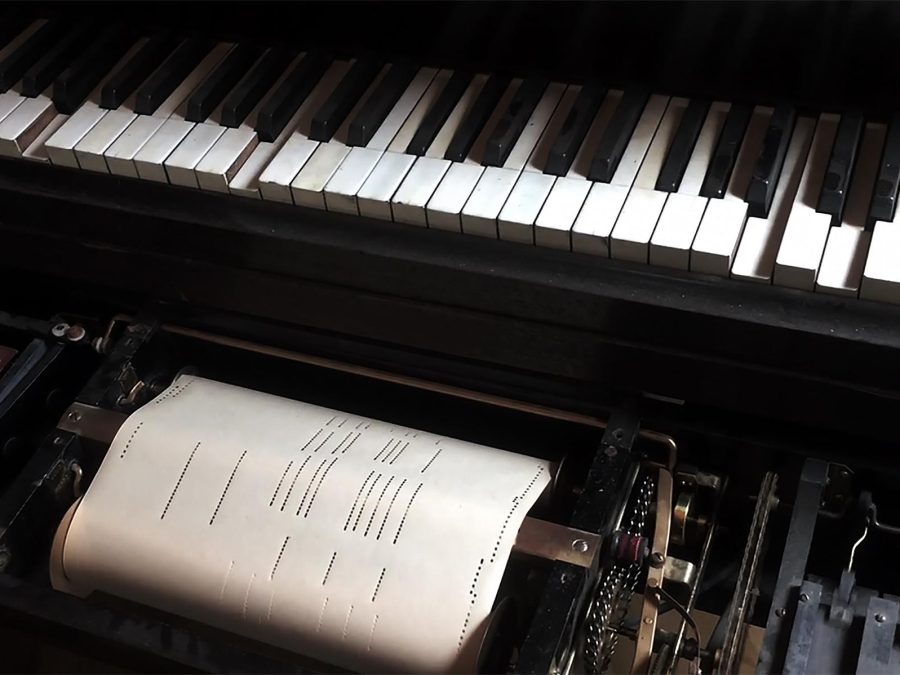 A close-up of a player piano keyboard captures the roll of sheet music mid-song.