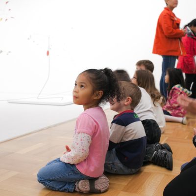 Little children sitting on a wooden floor in a gallery looking at Alexander Calder mobiles