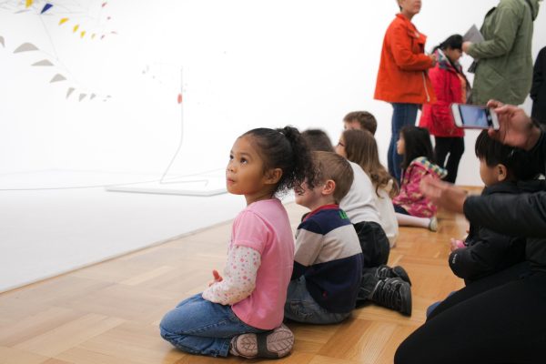 Little children sitting on a wooden floor in a gallery looking at Alexander Calder mobiles