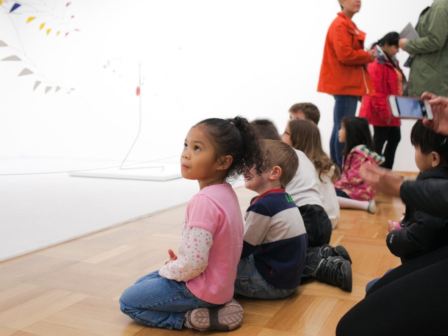 Little children sitting on a wooden floor in a gallery looking at Alexander Calder mobiles