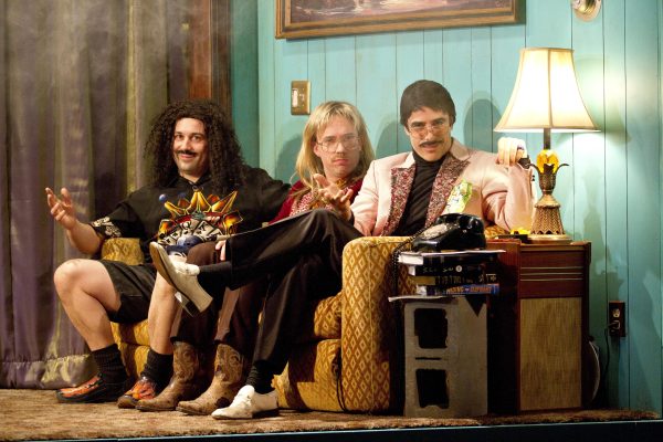Three people sit on a couch in a 1970s styled basement