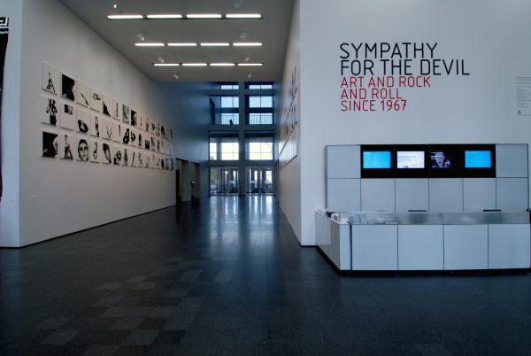 An el shaped two story room featuring a desk with tv monitors above it and text on the wall reading Sympathy for the Devil, Art and Rock and Roll Since 1967 in the foreground and an installation of paintings high up on the wall in the background
