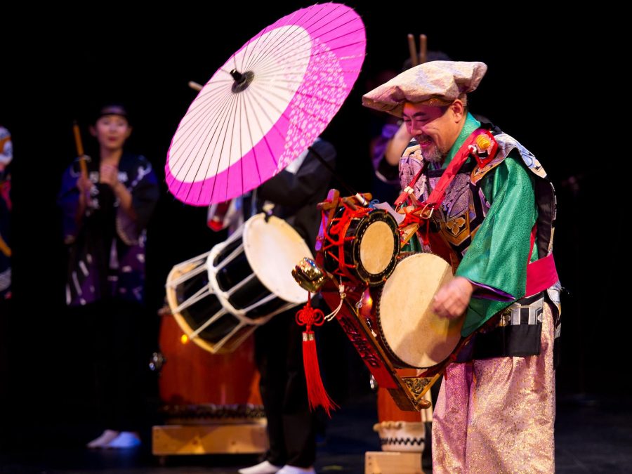 A group of people in colorful outfits, one with a pink washi parasol, carry various types of drums
