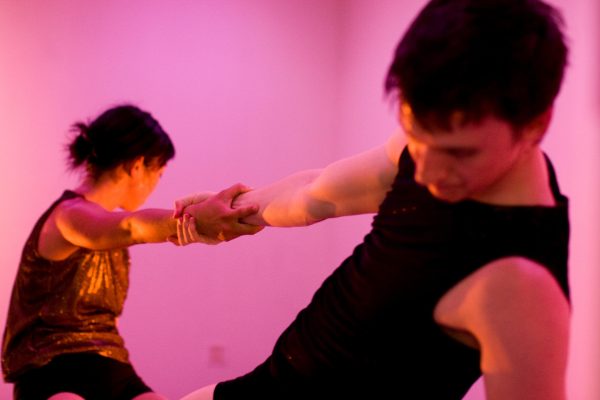 Two people lean away from each other, gripping the others right hand for balance, in a pink lit room