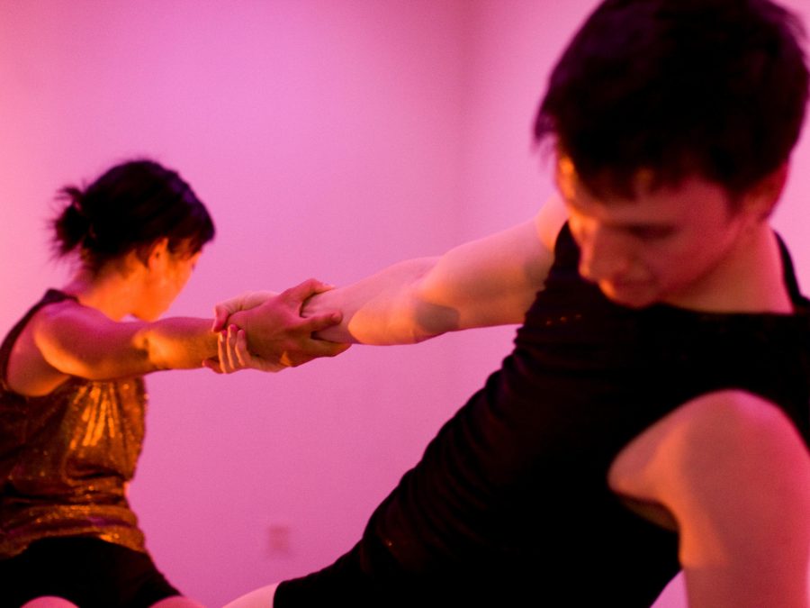 Two people lean away from each other, gripping the others right hand for balance, in a pink lit room