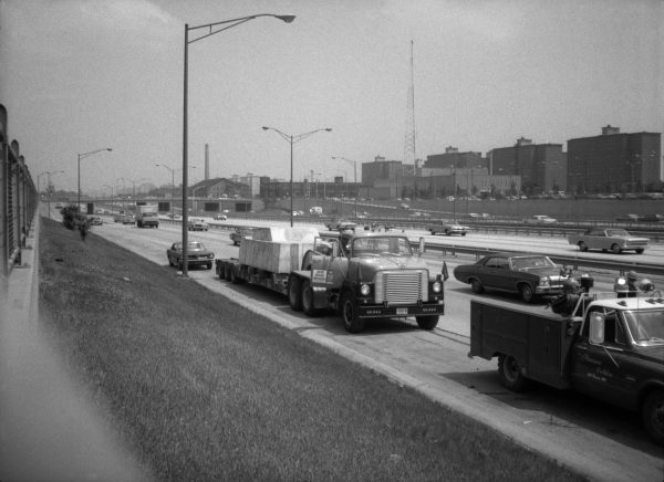 Vintage black and white photo of a busy highway scene with cars and trucks, including a prominent tow truck carrying a large concrete slab.