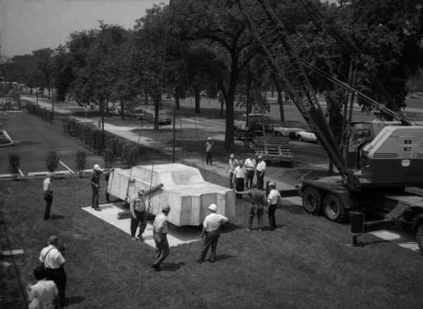A black-and-white photograph documenting a crew of people guiding a concrete car onto a base in the middle of a large park.