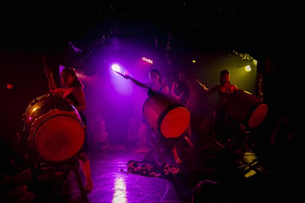 Three huge drums are held by women holding drumsticks high, they are illuminated in a dark space by pink, red, and yellow lights.