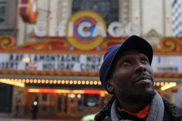 A young black man wearing a hat in front of the Chicago Theater looks up.