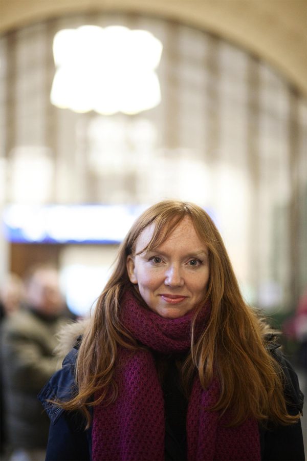 A pale-skinned person with long red hair wears a purple scarf and poses in front of a blurry background.