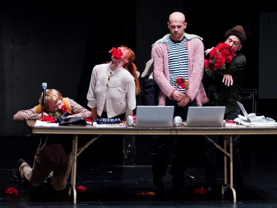 Three men and one woman stand or rest behind a rectangular, collapsible table with three laptops and various objects.Two people hold red flowers in their hands and arms; two have red flowers in their mouths.