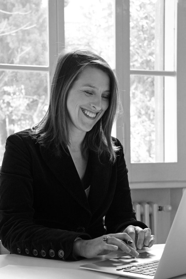 Black and white photograph of a female presenting person smiling down at a laptop computer in front of a three-paned window