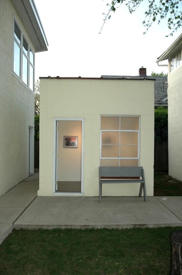 A small off-white building with a door and a window near other buildings