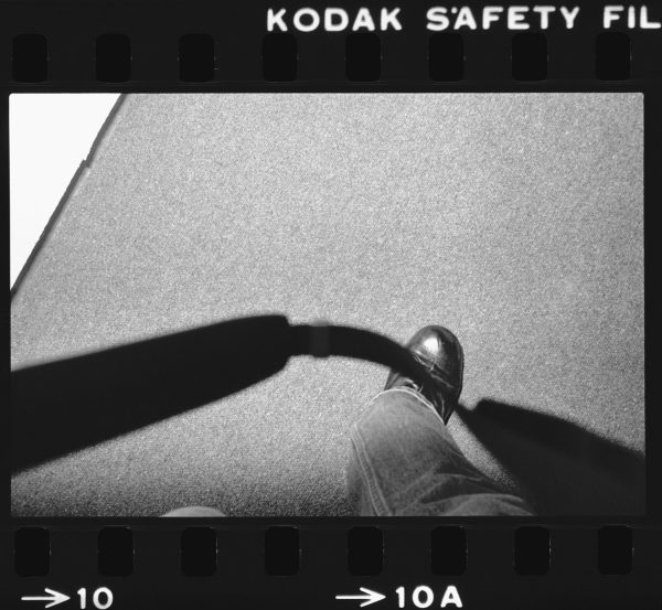 A 35 mm black-and-white film frame depicts a camera strap looping down toward the lower part of a right leg clad in denim as the person raises their foot to take a step.