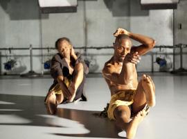 Two men sit on the gray floor of a dance studio. Each extends his right leg straight in front of them, with their left leg bent and held off the ground. The men gesture with their arms.