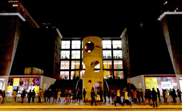 A line of people stand in front of a building featuring cutout sewer tubes decorating a large plaza