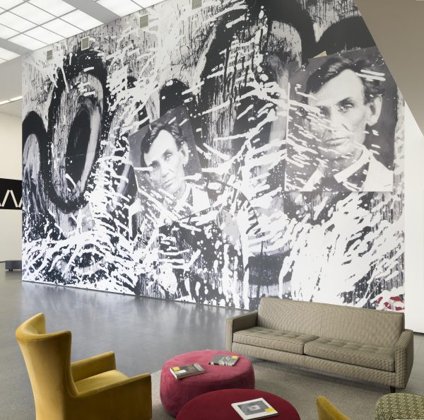 An image of a huge photographic wallpaper mural in the MCA's second floor lobby featuring cartoonish eyes, two photographs of Abraham Lincoln, and splatters of black and white paint. A setting of living room furniture is in the foreground of the image.