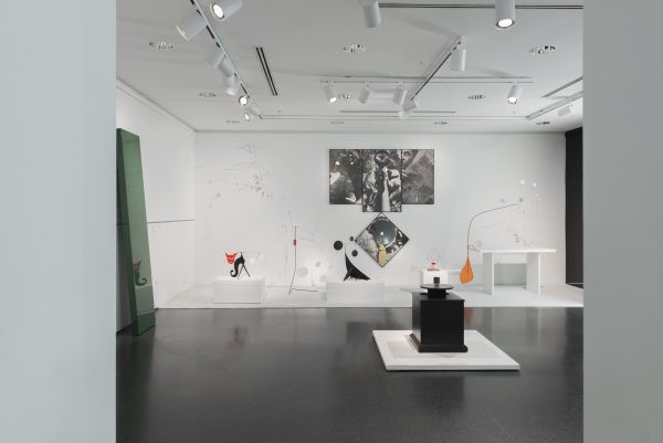 Several mobile sculptures in various shapes rest on pedestals or hang from the ceiling in front of a large multipaneled black-and-white photograph in a white-walled gallery.
