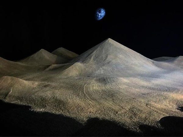 In a dark room, a large mound of sand forms several cone shapes, while a small light projection in the background shows an abstract, vaporous shape that resembles a planet.