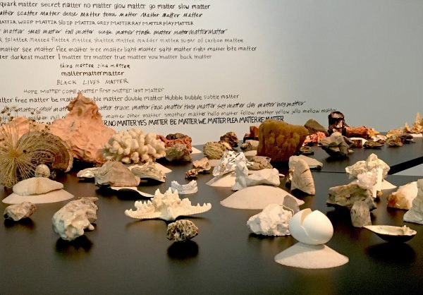 A table with rocks, minerals, and other substances sits in front of a wall covered with black text.