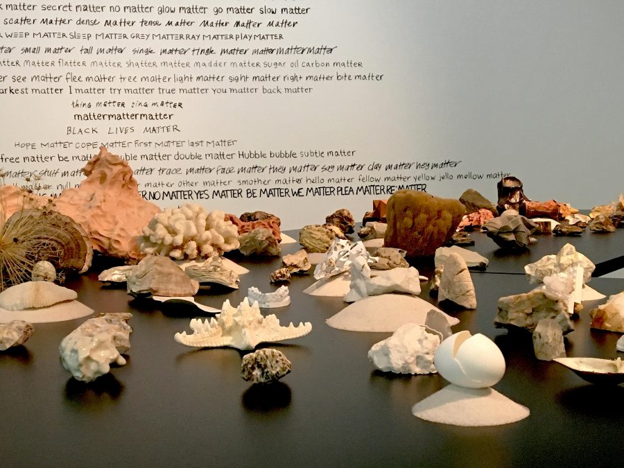 A table with rocks, minerals, and other substances sits in front of a wall covered with black text.