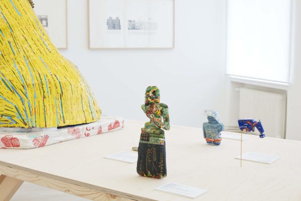 A little figurine made out of colorful packaging on a wooden table next to other objects with drawings hanging on the wall behind it