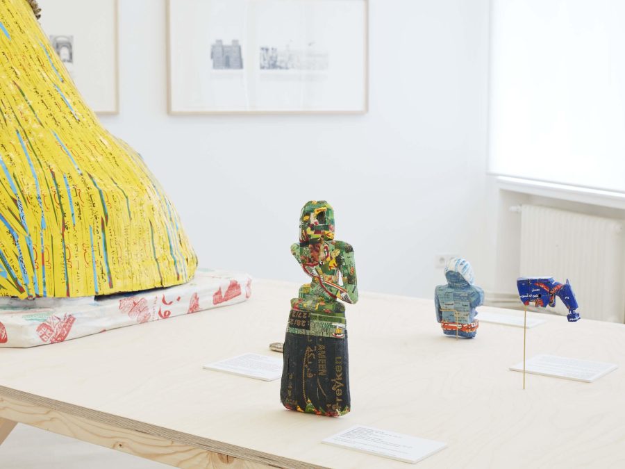 A little figurine made out of colorful packaging on a wooden table next to other objects with drawings hanging on the wall behind it