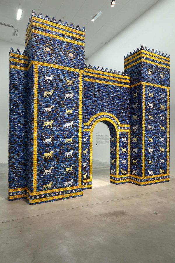 An architectural arched gateway is shown, installed within a modern gallery space. The arch is made from bricks in a range of different blues with golden yellow accents and decorated with stylized animals.