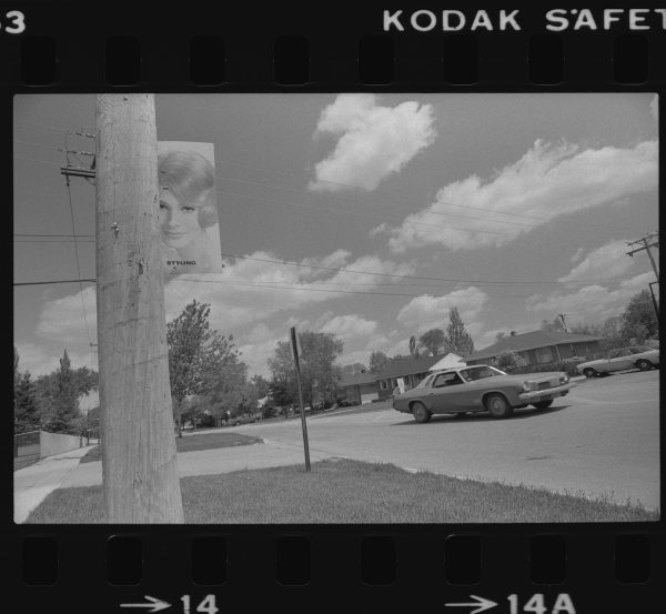 A black-and-white film frame shows a car driving along a suburban street. In the foreground is a wooden utility pole affixed with a poster of a woman and the the word 