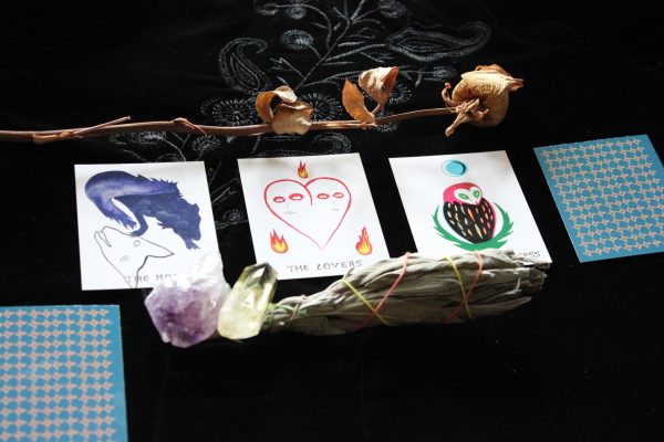Four tarot cards are placed in a row on an embroidered black tablecloth. Three of the four cards reveal an image. A dried rose frames the top of the photo. Two crystals and a smudging stick are in the foreground. A fifth unrevealed card is cut off in the lower left corner.