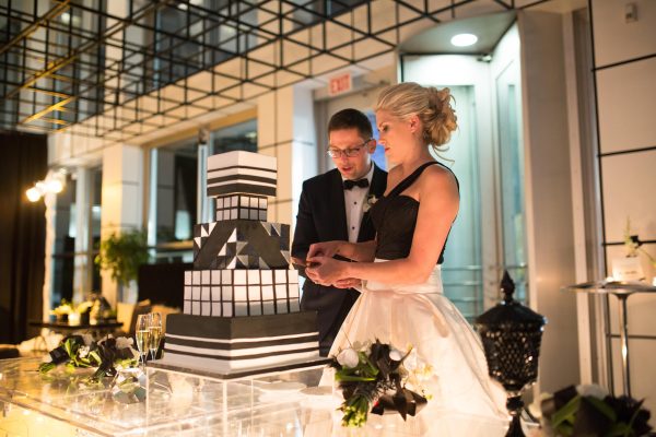 A bride and groom cut a black and white cake in a room featuring a black, 3D grid on the ceiling