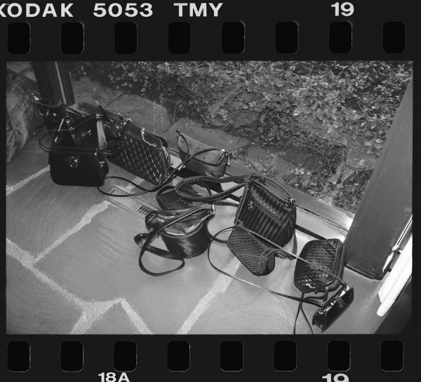 This black-and-white 35 mm film frame depicts nine purses of varying shapes and sizes on a stone floor next to a glass window.