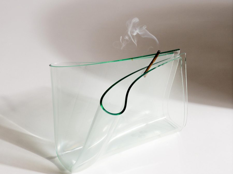 A piece of glass is folded into quarters with green edges facing up, resting on a white surface. A smoking stick is wedged into the top fold. There is a dark, round shadow behind the sculpture on the right.
