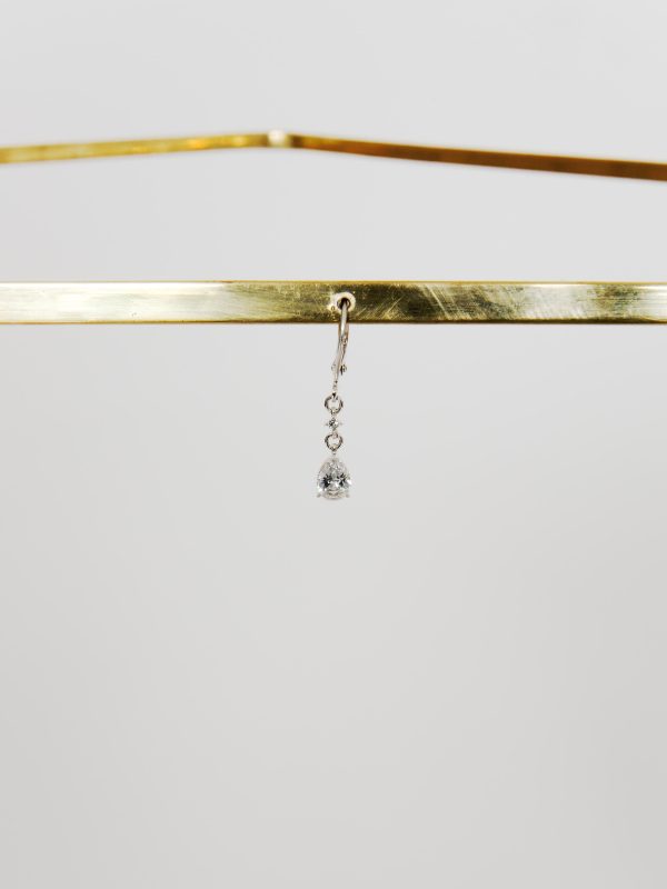 A close up image of a bronze-colored strip of metal cutting horizontally across the frame, with a single diamond earring dangling from its center. Another bent piece of similar metal is shown out of focus above and behind it.