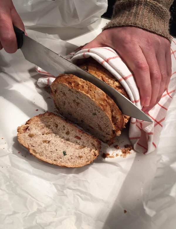 A person cuts a second slice of bread while holding the rest of the loaf with a towel