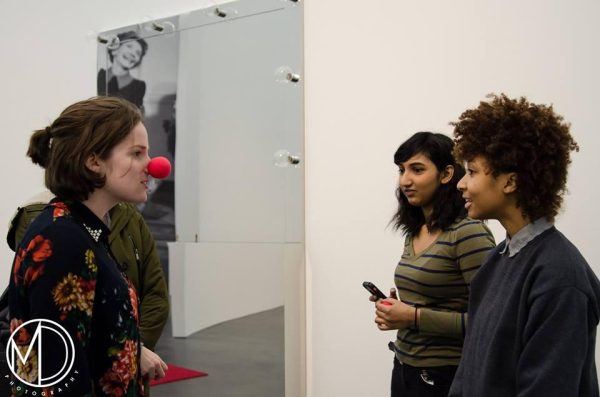 Three teenager girls stand talking together near a mirrored artwork; one wears a red Bozo-the-Clown nose. A watermark on the bottom left reads: 