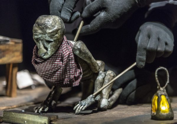 A grimy puppet kneels on a wooden surface, a checkered cloth wrapped around its neck, and a model lantern glowing yellow beside it. The puppet is operated by sticks, held by four black-gloved hands.