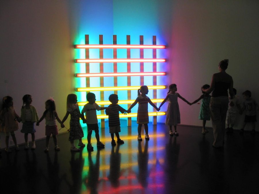 A line of children and an adult are cast in a shadow as the stand in a dark room next to a neon light gird formation