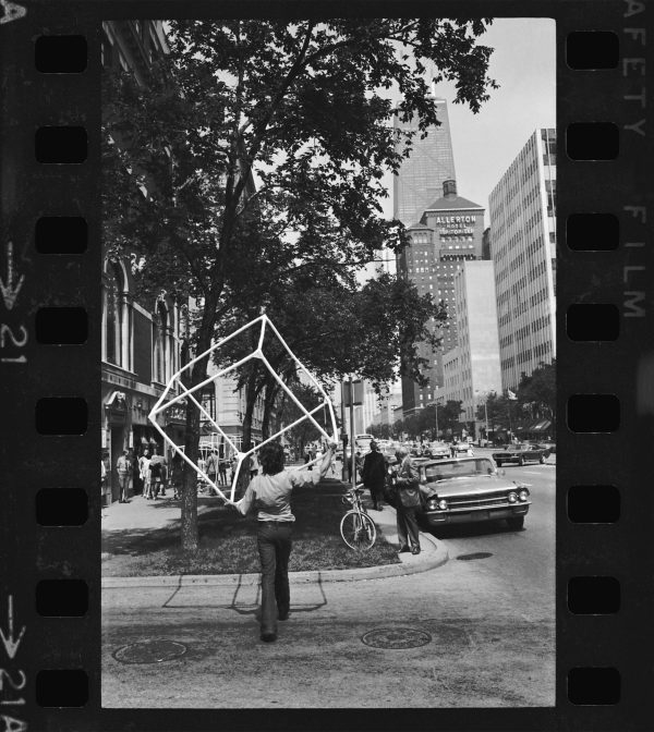 A black-and-white film frame shows a woman carrying an open cube sculpture while crossing the street, heading North on Michigan Avenue, Chicago, with the Allerton and John Hancock buildings in the background.