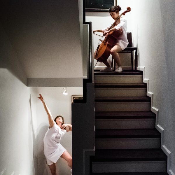 There are two women in a stairwell performing: one on the upper landing plays a cello and looks at the camera while the other woman appears on a lower level, lifting an arm and leg up as if dancing.