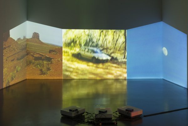 Three projectors on the floor project three different images on the opposite wall: a southwestern desert landscape, a car in golden light, and a small moon on a blue sky. The images are projected on the corners of the room, creating irregular viewing frames.