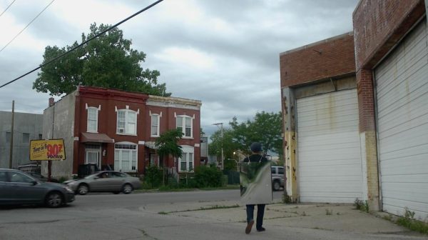 On a cloudy day, a man with his back to the camera, wearing a walking sandwich board sign made of reflective material stands in an urban outdoor environment surrounded by an old garage, some cars, trees and brick buildings.