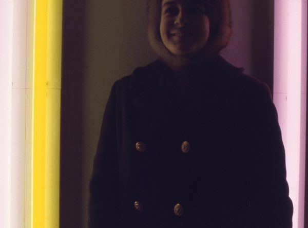 A person in a winter coat smiles near vertical neon lights