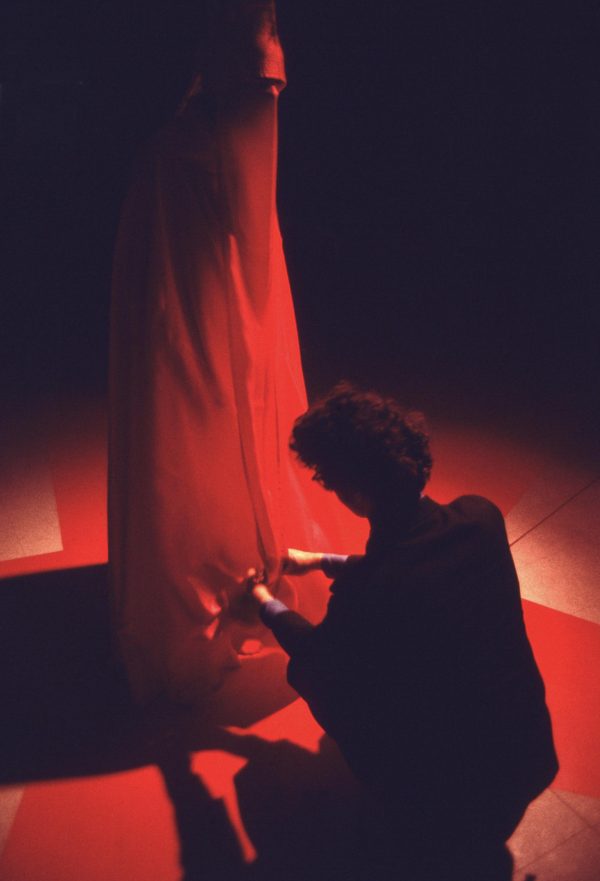 A person squats facing away engaged with an red fabric abstract sculpture lit by a spotlight in an otherwise dark room