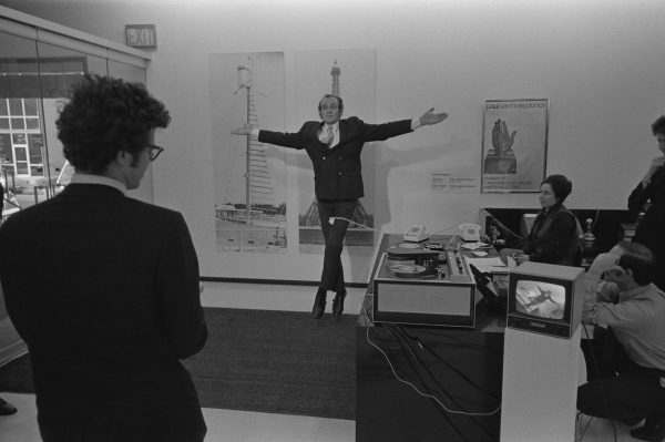 Archival photo of a man in a suit performing a sauté ballet jump as other people look on. His movement is simultaneously projected on a small television.