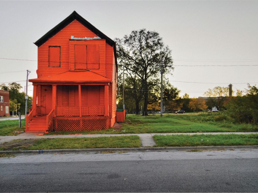 A bright red-orange house is boarded up, standing next to an empty urban lot during sunset.