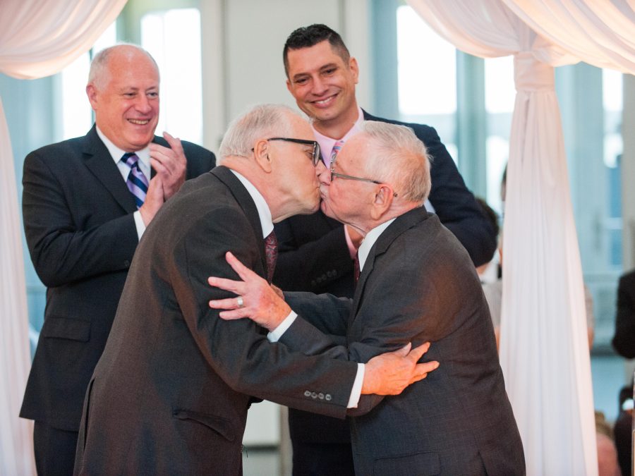 Two elderly men wearing suits embrace in a kiss while two other men look on smiling and clapping.